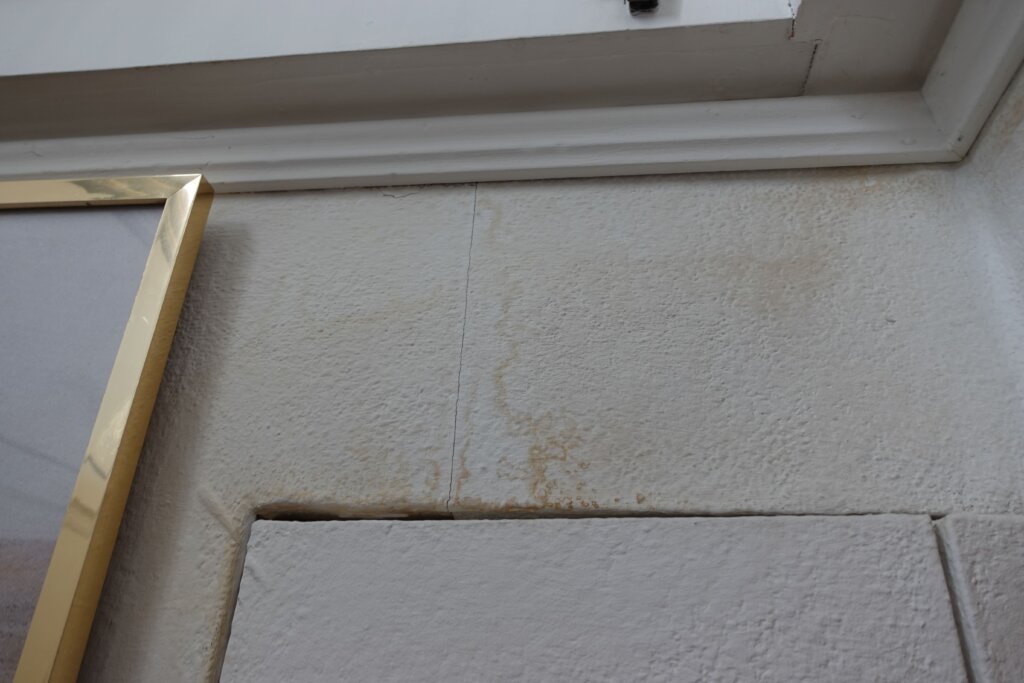 Nahaufnahme einer Wandecke mit weißen Fliesen, Holzleiste und sichtbaren Fugen sowie leichten Verfärbungen