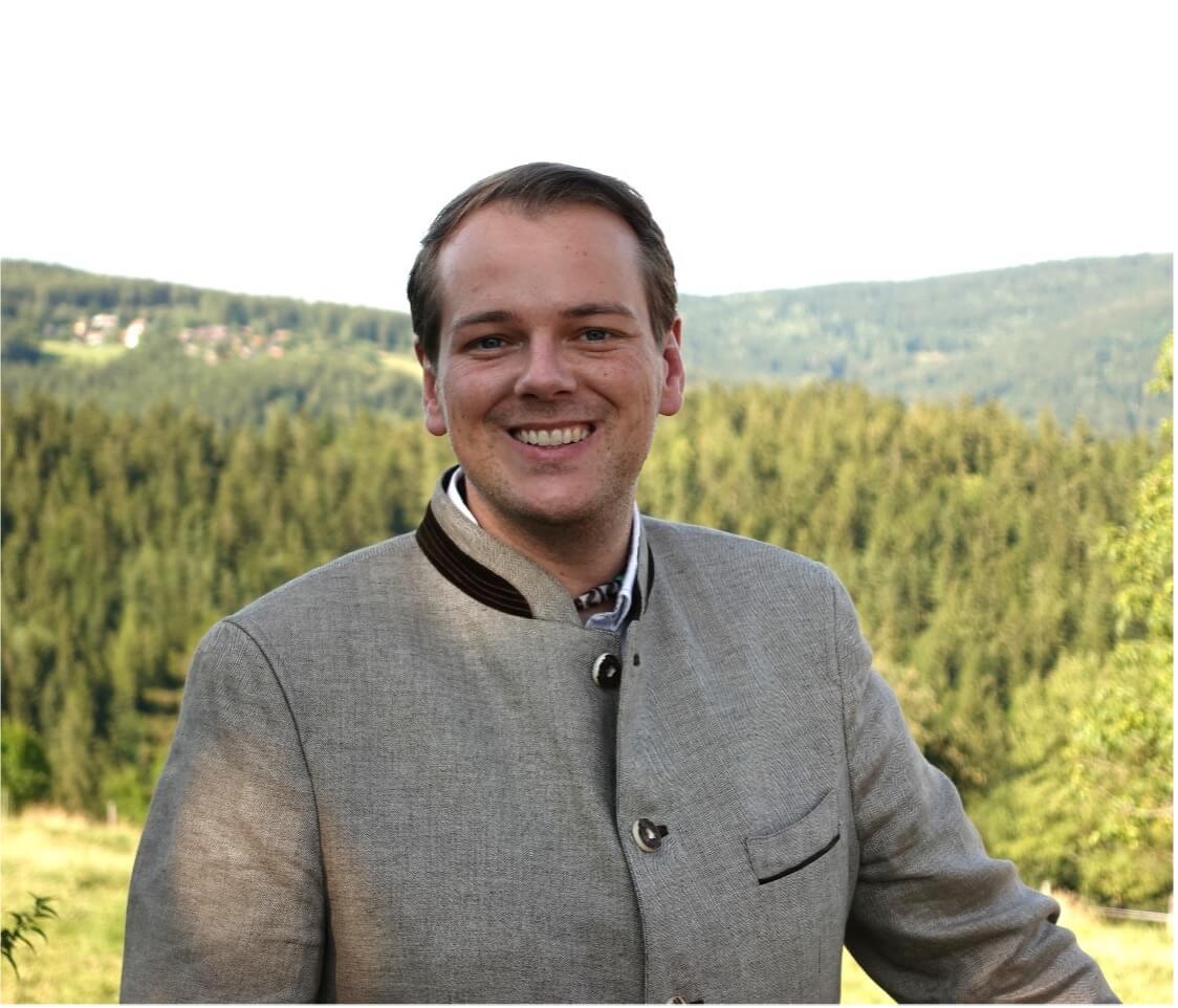 Mann mit grauem Jacket lächelt vor bewaldeter Berglandschaft